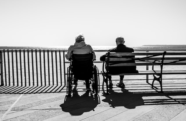 photo of the back of older couple sitting looking at the ocean, one on a bench, one in a wheelchair
