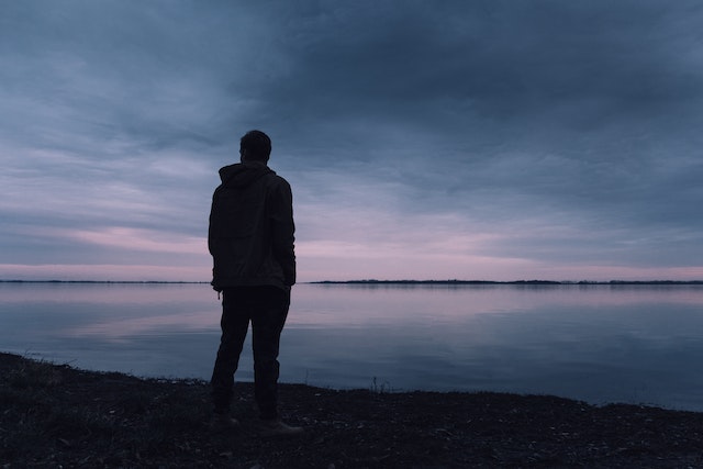 photograph of a silhouette of a man standing at the edge of a body of water with a dusk light